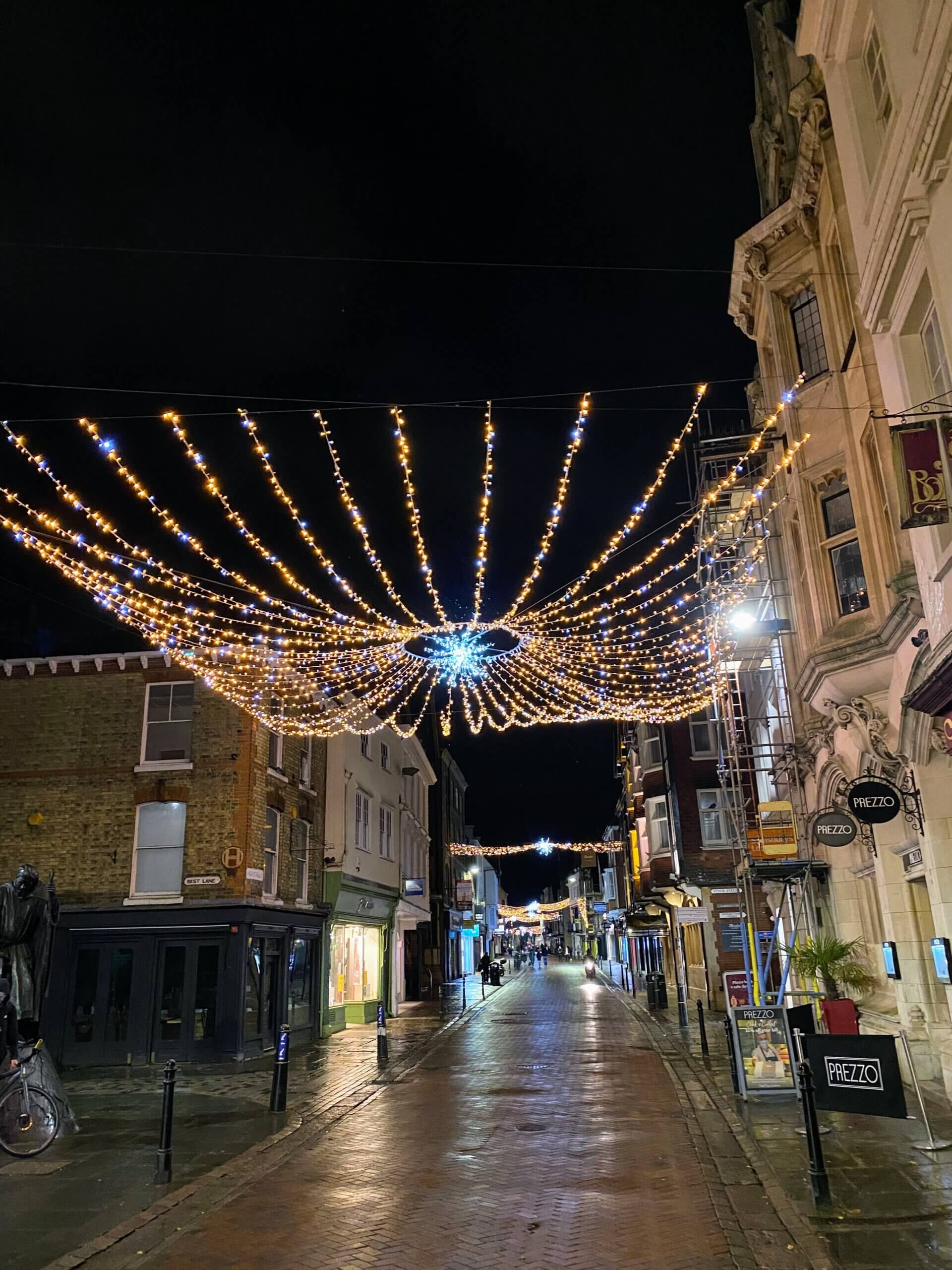 A charming street in Canterbury illuminated by festive lights hanging overhead, creating a warm and inviting atmosphere at night.