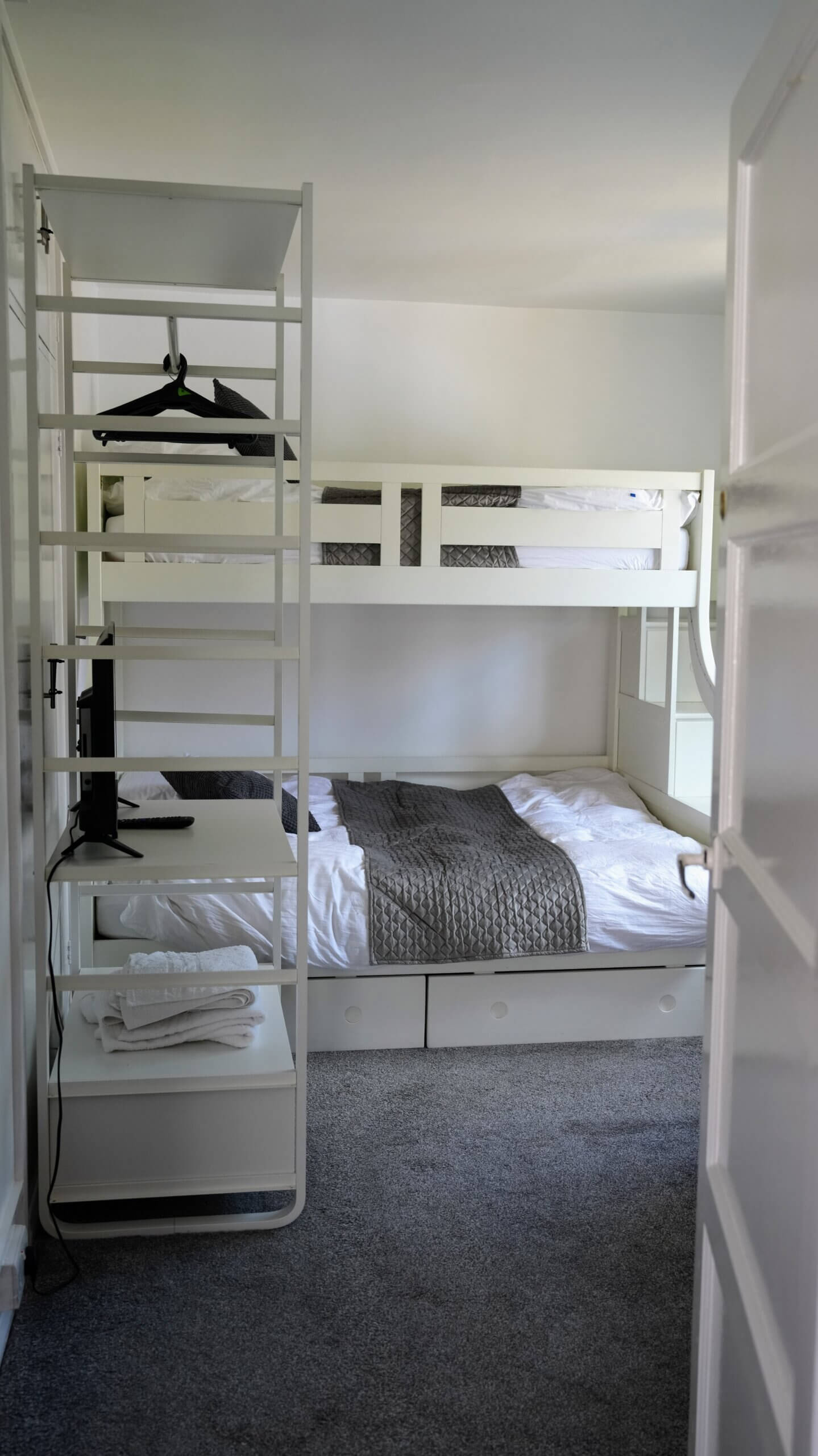 A bright and tidy bedroom featuring white bunk beds with neatly made bedding, built-in storage drawers, and soft grey carpet.